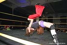 A Caucasian female wrestler executes a throw that involves lifting her opponent up in the air and slamming the opponent's back inside a wrestling ring.