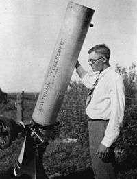A young man in his mid-twenties, wearing glasses, a white shirt, tie and long trousers, stands in an open field, next to a Newtonian telescope resting on the ground and tilted towards the sky. The telescope is taller than him, and is about eight inches in diameter. His right hand is resting up on the barrel, and he looks slightly past the telescope, out to the left.