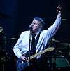 A man wearing a black tie and white dress shirt with an electric guitar hanging from a strap around his neck. His left arm is raised, with his index finger pointed upwards. In the background is a drum set.