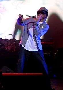 Siyah pantolon, beyaz penye ve gri kapşonlu giymiş bir rapçi, kapşonunun şapkası kapalı bir şekilde elindeki mikrofona şarkıyı söylüyor.