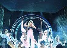 A blond woman in white pants and black bustier stands inside a number of metallic circular rings. She holds a microphone to her mouth with her left hand and her right hand makes a claw like gesture. The woman is flanked by men and women in white dress and fierce expressions with their eyes being white in color.