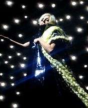 A blond woman wearing a shiny black dress and her hair extended with long braids, stands on a stage. The braids fall on her left arm and continue down. Behind her, a number of twinkling lights are visible and the woman makes a gesture with her right hand.